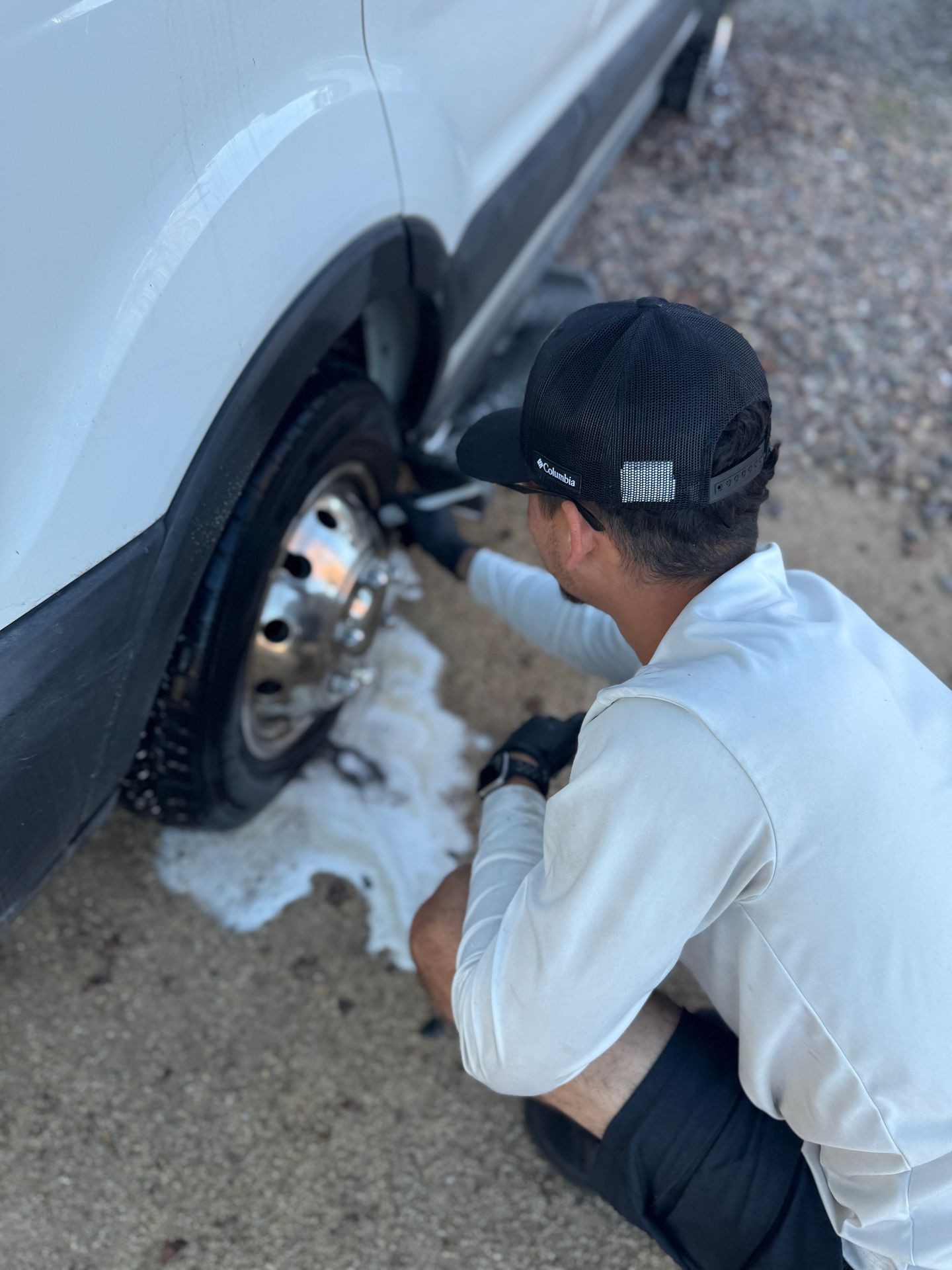 Man cleaning a car wheel with white foam, wearing a black cap and gloves.