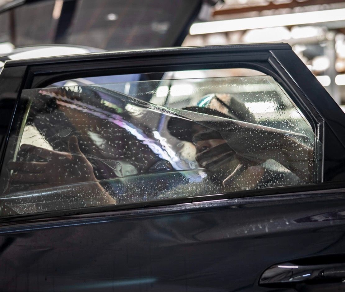 Person installing window tint on a car door, visible through the glass with water droplets.