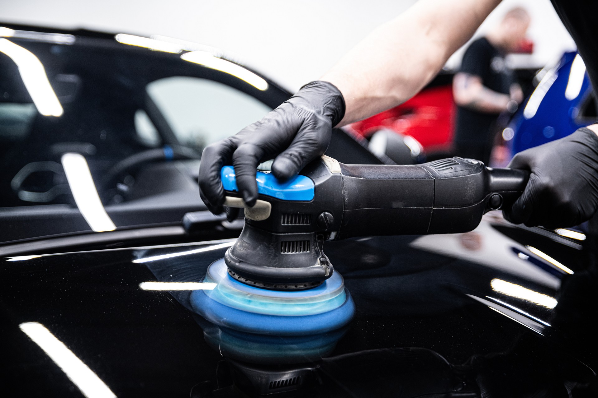 detailer wearing black gloves polishing car paint with a machine in a detailing studio.